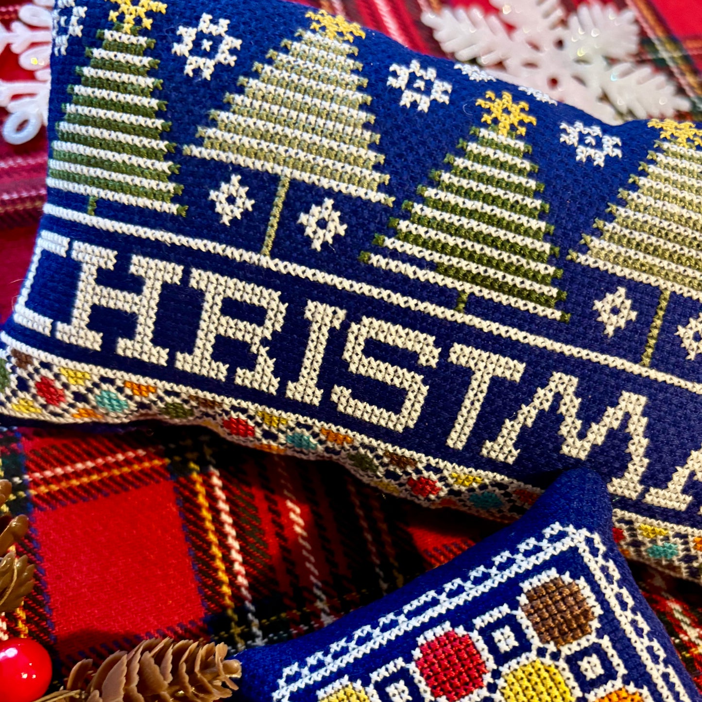 Cross-stitched Christmas-themed pillow with trees and snowflakes on a plaid fabric background.