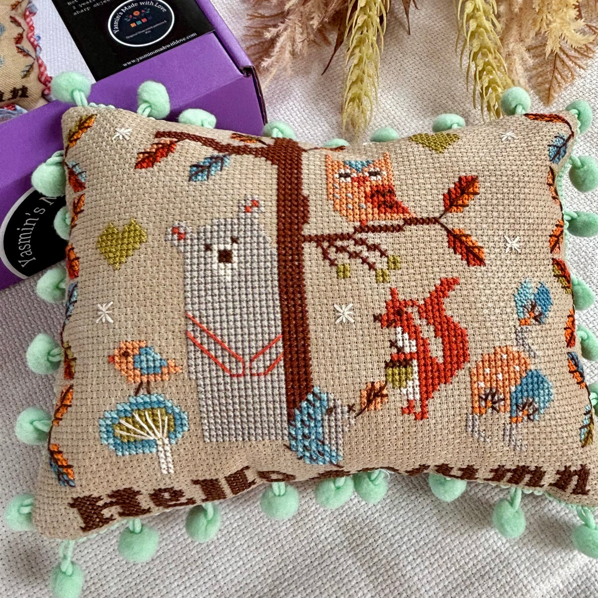 Decorative pillow with embroidered animals and 'Hello Autumn' text, surrounded by green pom-poms.