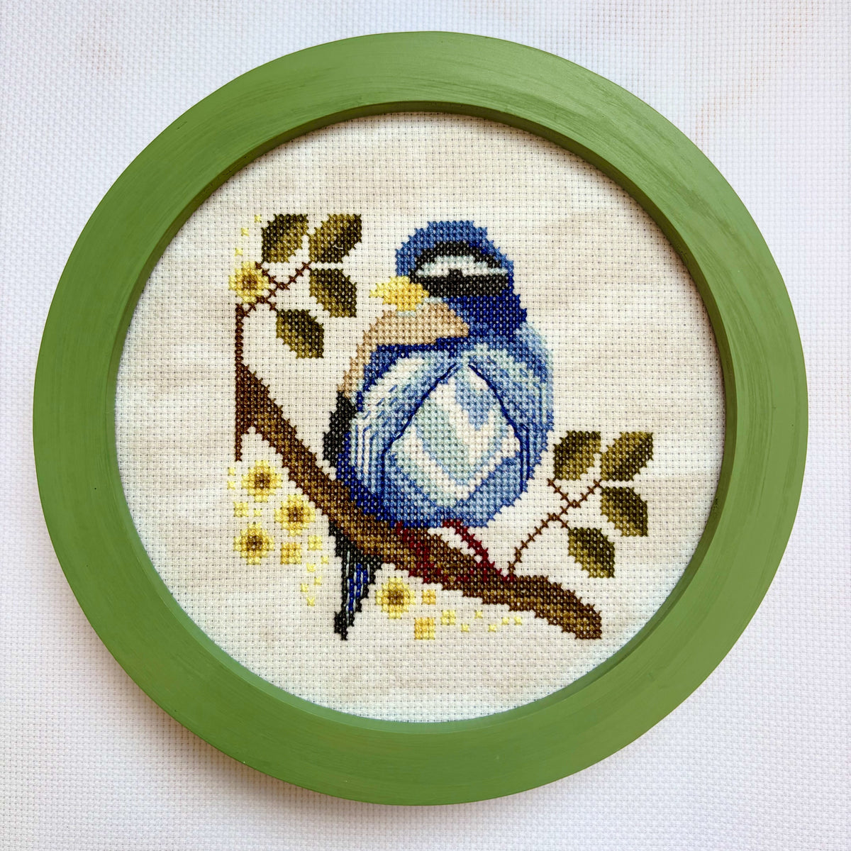 Cross-stitched blue bird on a branch with yellow flowers in a green frame.