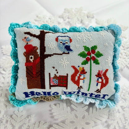 Decorative pillow with cross-stitched design featuring animals and 'Hello Winter' text on a textured surface.