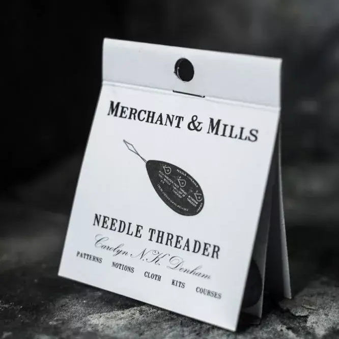 A white paper card package for a Merchant & Mills needle threader, photographed standing on a dark textured surface with a dark blurred background.