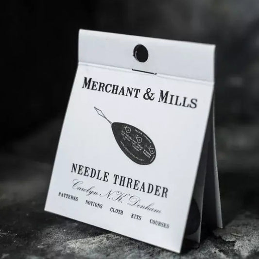 A white paper card package for a Merchant & Mills needle threader, photographed standing on a dark textured surface with a dark blurred background.