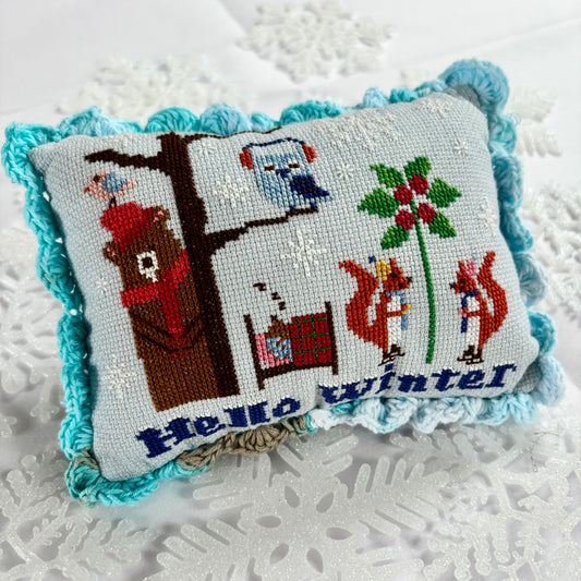 Decorative pillow with cross-stitched design featuring winter scenes and text 'Hello winter' on a textured white background.