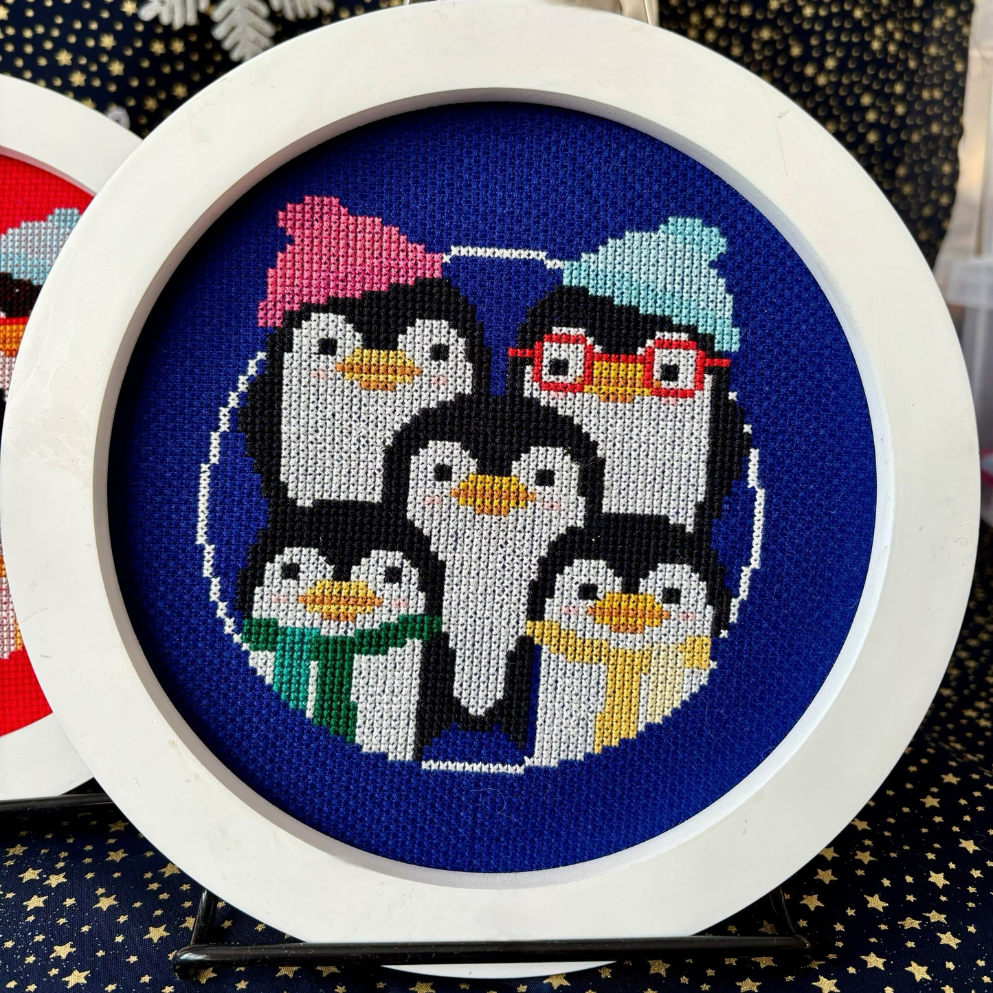 Cross-stitched penguins with colorful hats and scarves in a white hoop on a blue fabric background.