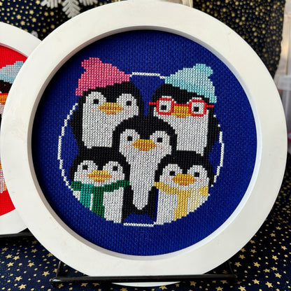 Cross-stitched penguins with colorful hats and scarves in a white hoop on a blue fabric background.