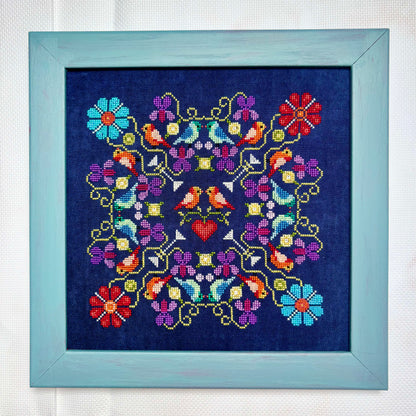 Framed cross-stitched pattern with colorful flowers and birds on a blue background