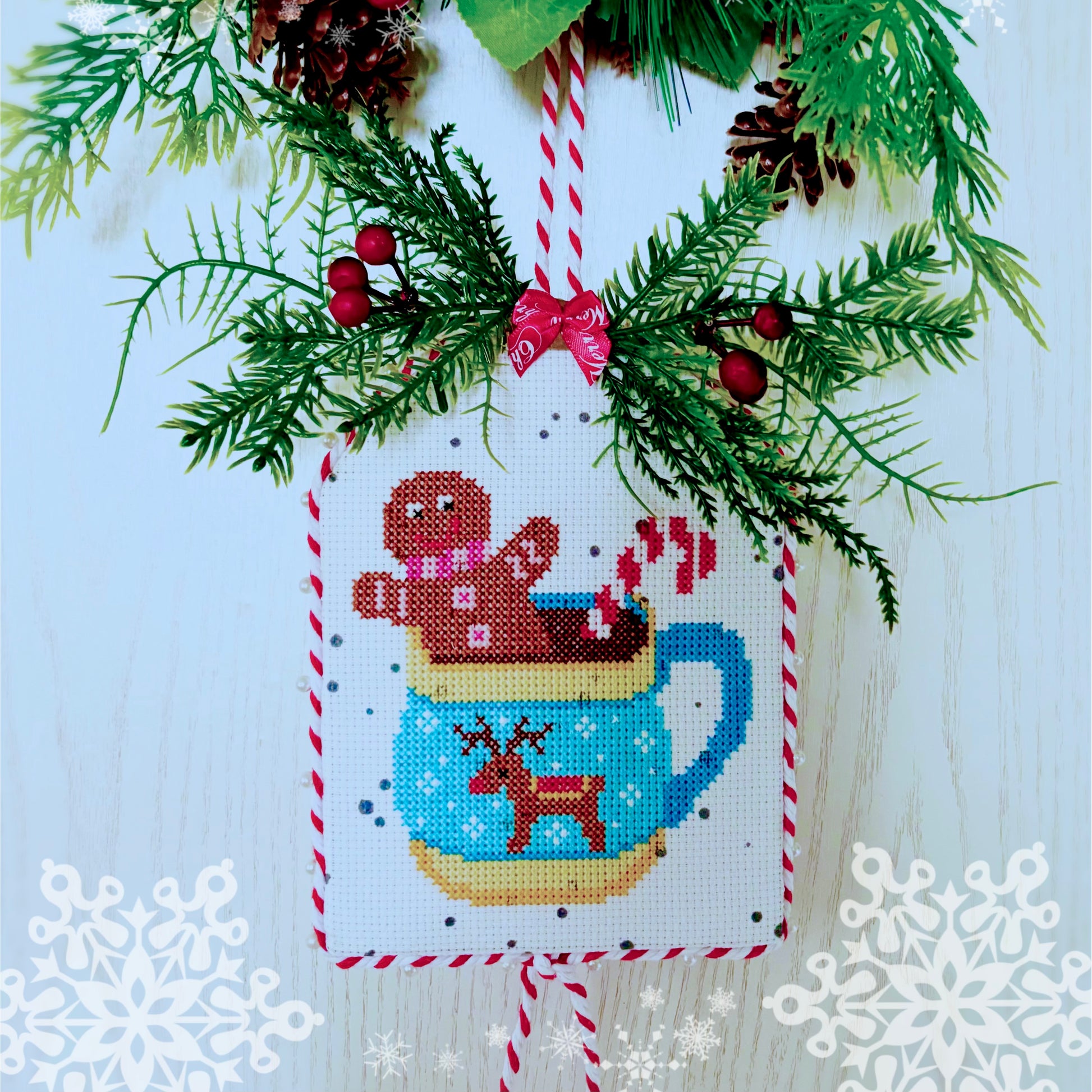 Cross-stitched design of a Christmas-themed cup with gingerbread man and reindeer, surrounded by festive elements on a light blue background.