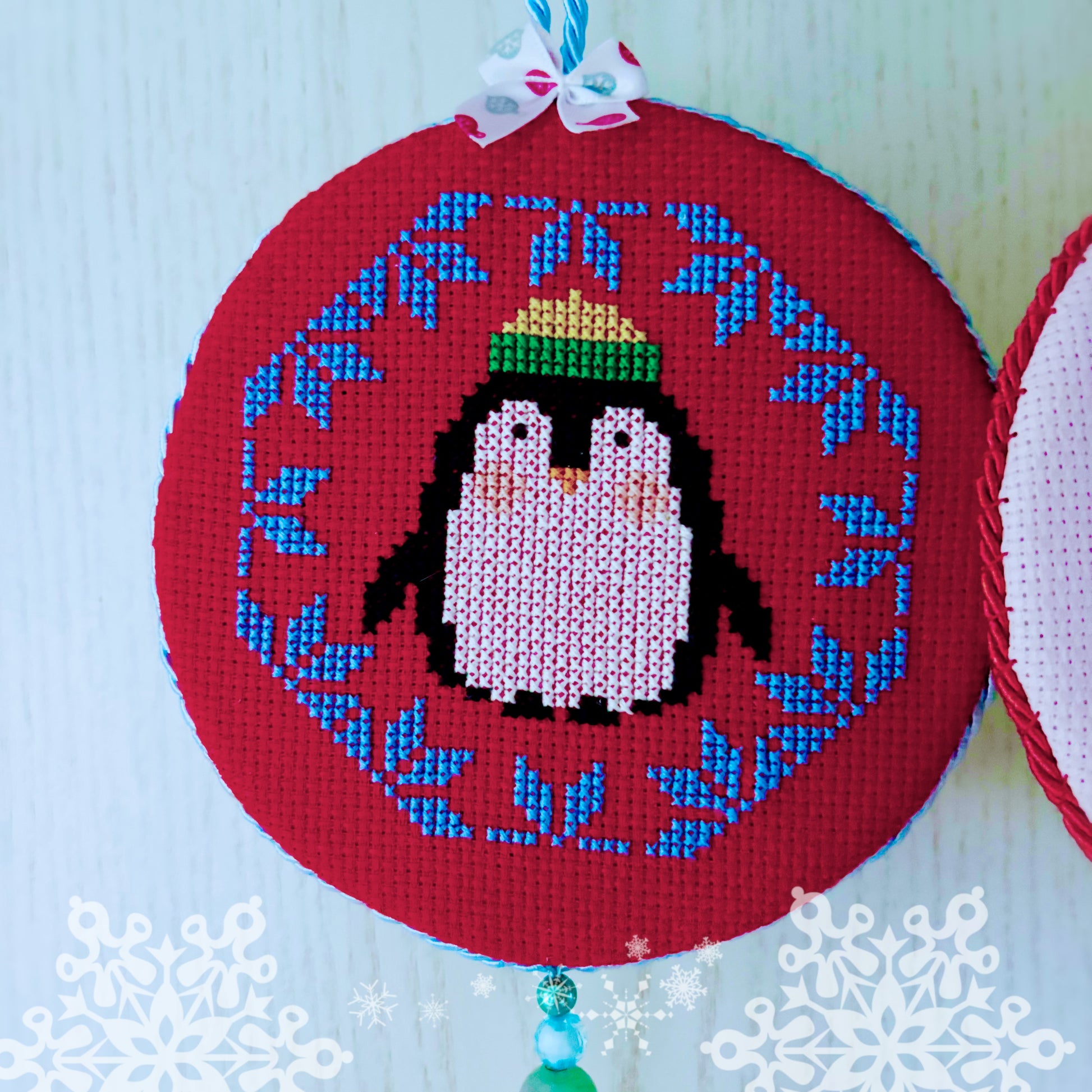 Cross-stitched penguin design on a red hoop with blue floral patterns, against a light blue background.