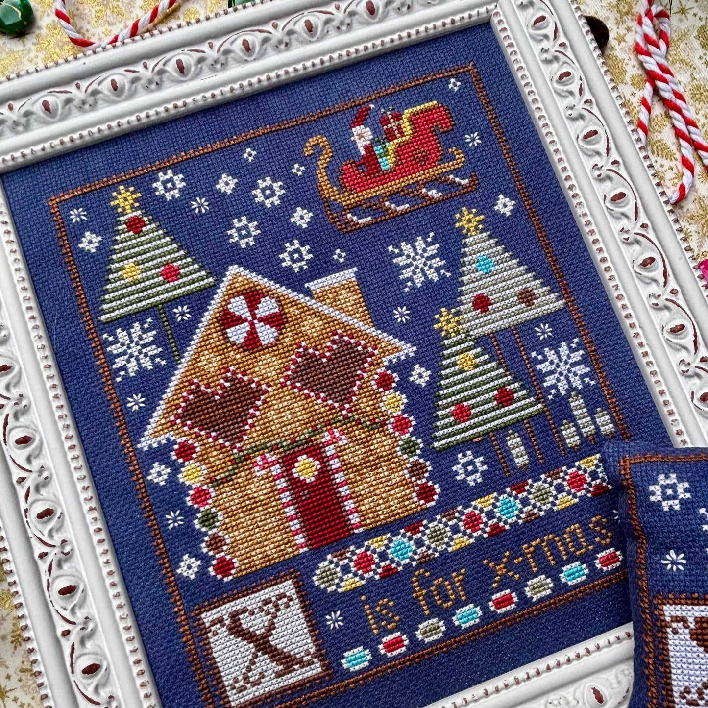 Cross-stitched Christmas scene with gingerbread house, trees, and 'X is for Christmas' text on a blue background.
