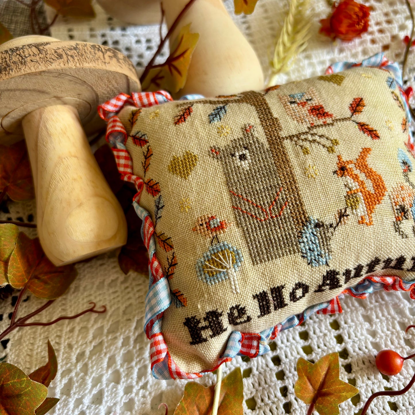 Handmade cross stitch pillow with woodland animals, colorful autumn leaves, and the text "Hello Autumn," stitched on linen and surrounded by fall decor