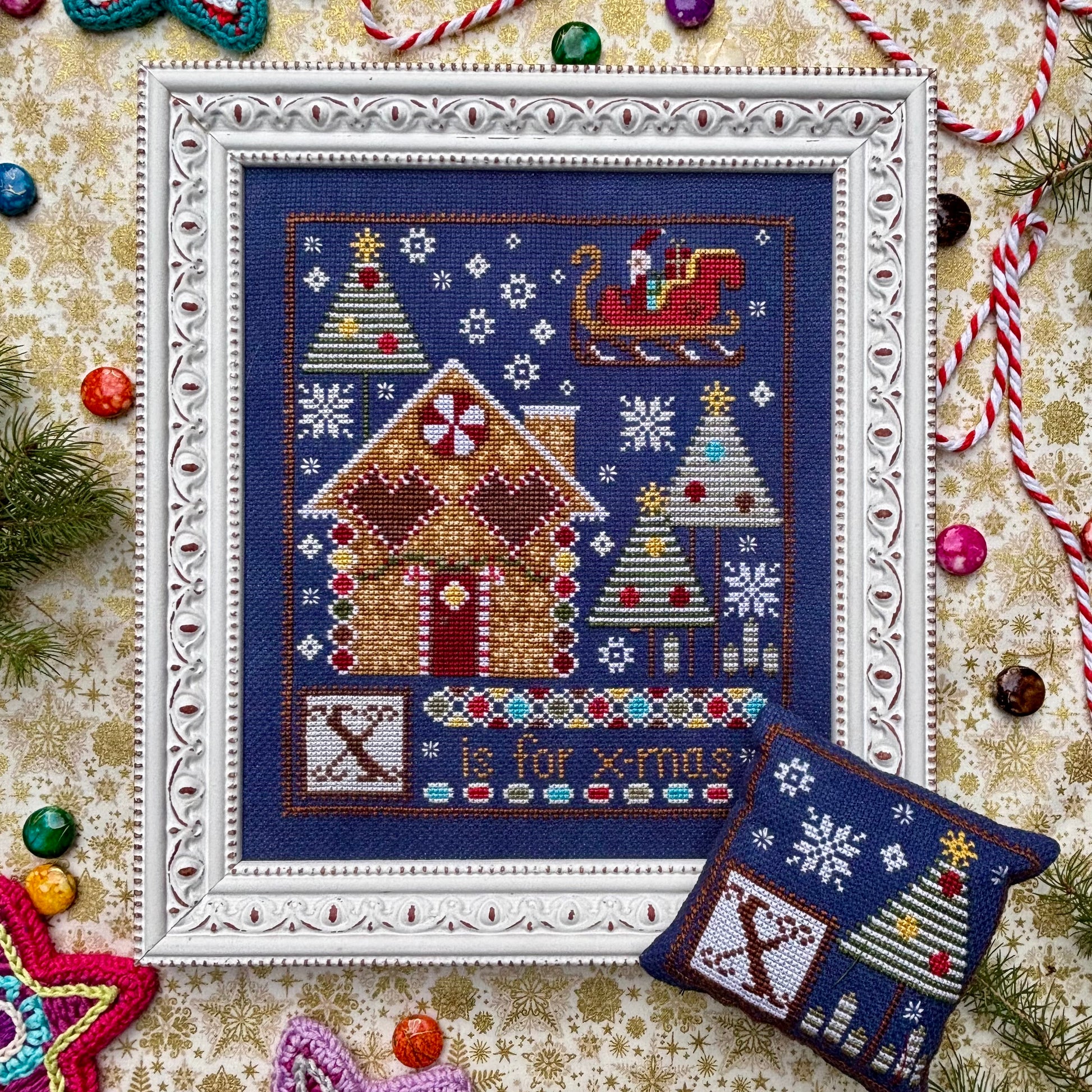 Cross-stitched Christmas scene with gingerbread house and trees on a decorative background.