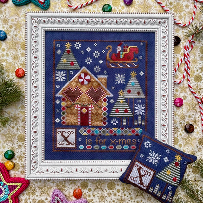 Cross-stitched Christmas scene with gingerbread house and trees on a decorative background.
