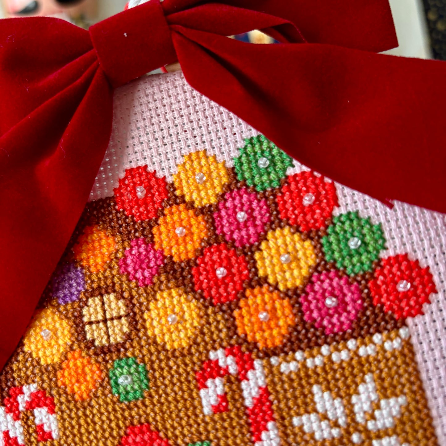 Cross-stitched gingerbread house with colorful candies and a red bow on top.