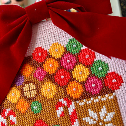 Cross-stitched gingerbread house with colorful candies and a red bow on top.