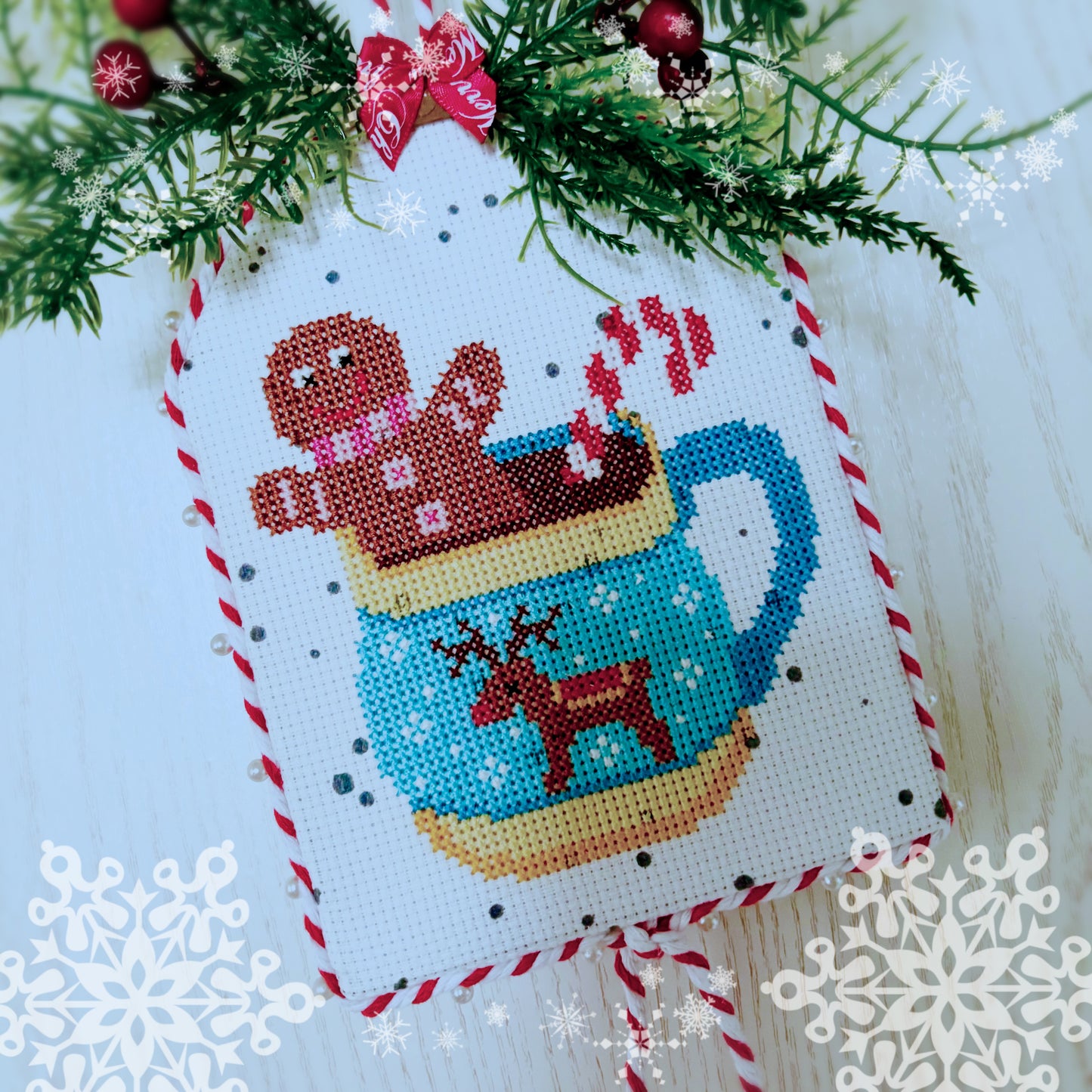 Cross-stitched gingerbread man in a mug ornament on a Christmas tree with snowflakes.