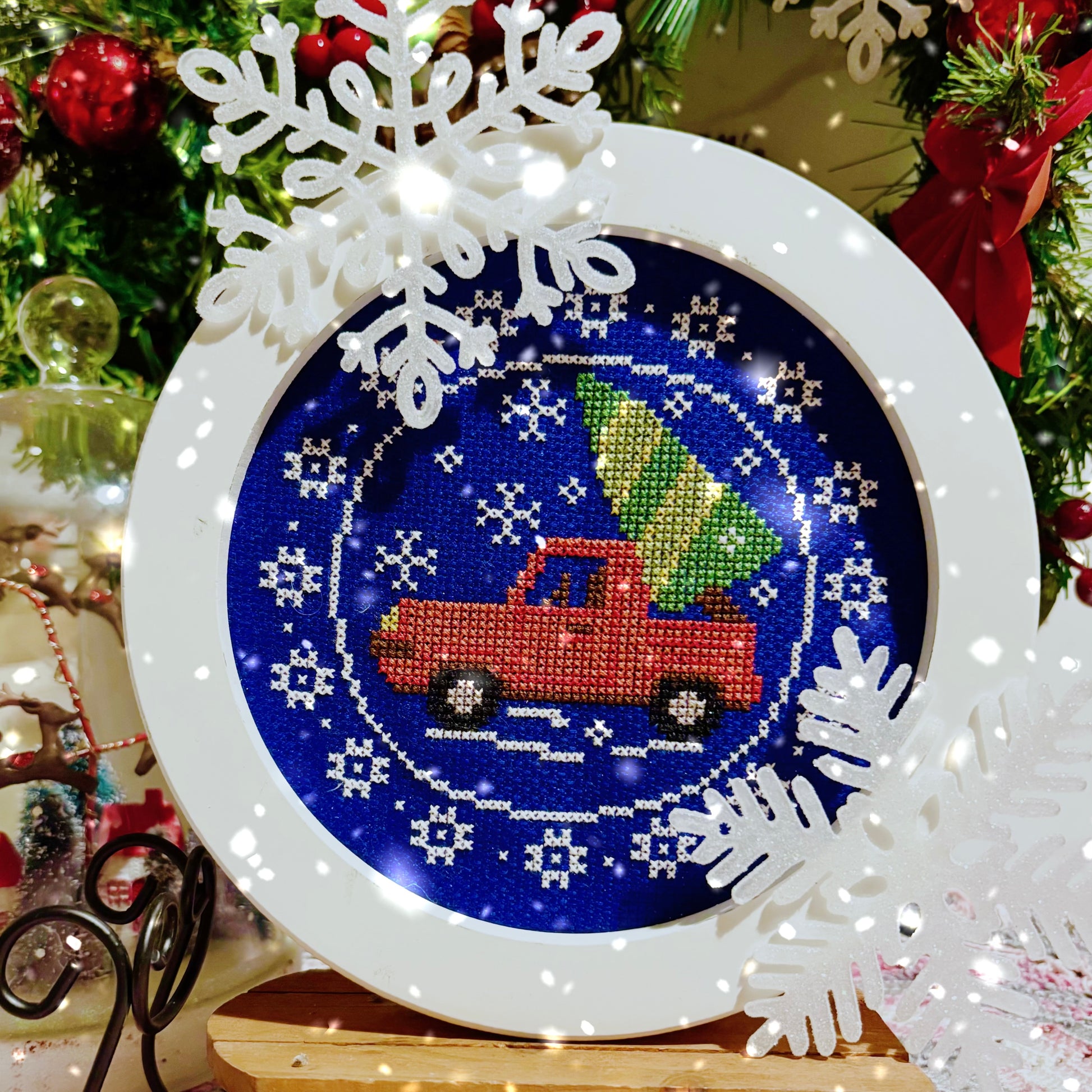 Decorative cross-stitch design with a red truck and Christmas tree design, surrounded by snowflakes and a festive background.