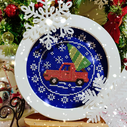 Decorative cross-stitch design with a red truck and Christmas tree design, surrounded by snowflakes and a festive background.