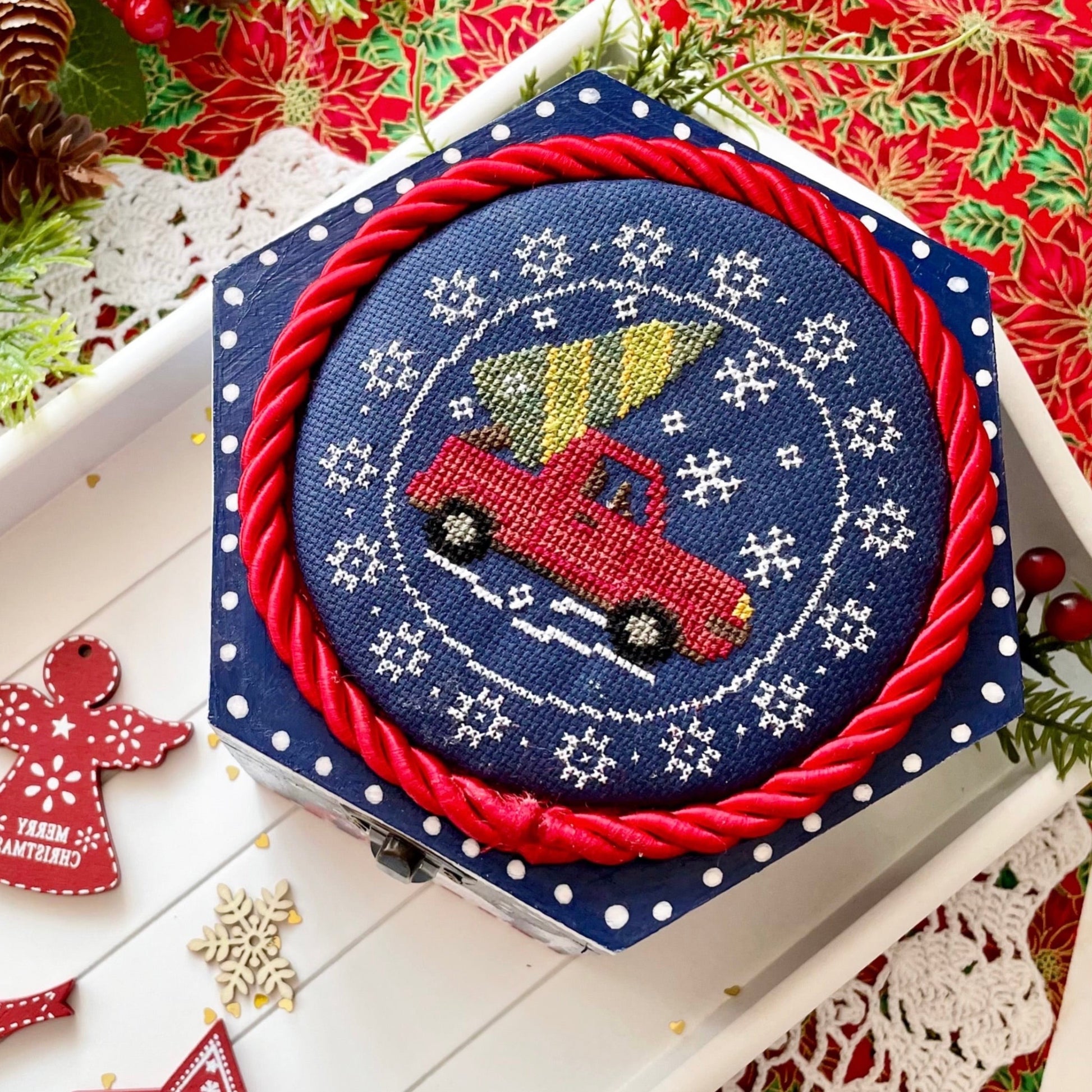Cross stitch kit with red truck and Christmas tree design on a blue background, surrounded by festive decorations.
