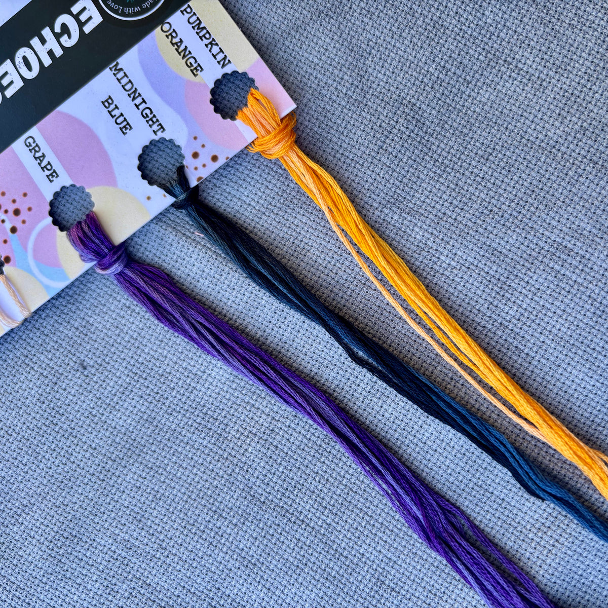 Three skeins of embroidery floss in Grape purple, Midnight blue, and Pumpkin orange, displayed on a gray textured fabric background with their packaging card.