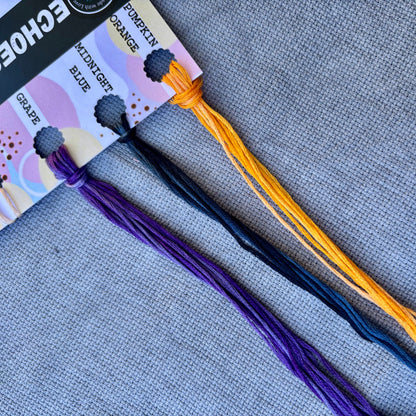 Three skeins of embroidery floss in Grape purple, Midnight blue, and Pumpkin orange, displayed on a gray textured fabric background with their packaging card.