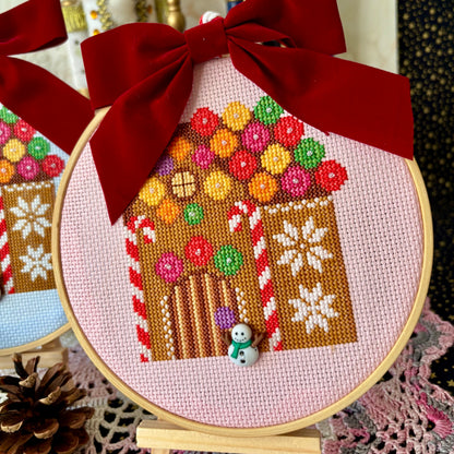 Cross-stitched gingerbread house in an embroidery hoop with a red bow, surrounded by festive decorations.