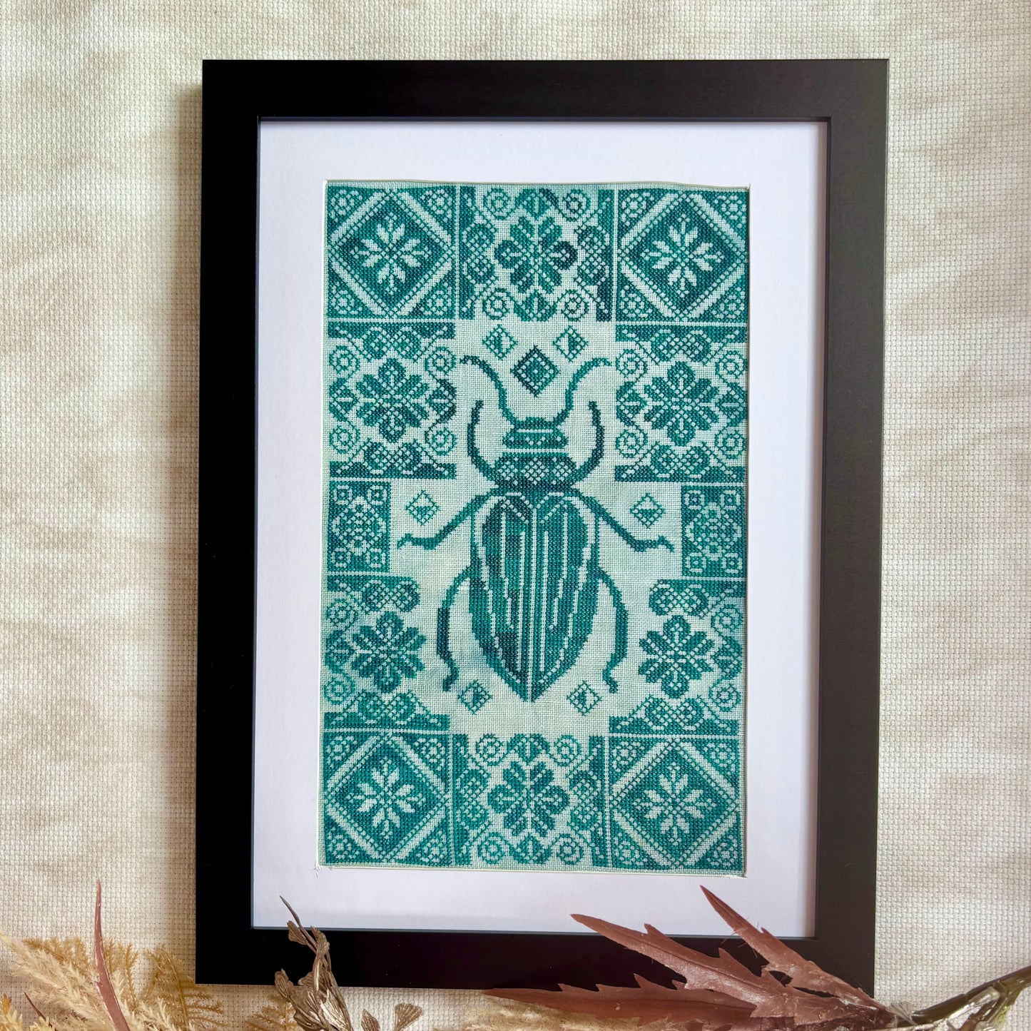 Framed cross-stitched pattern with a beetle design on a textured wall.
