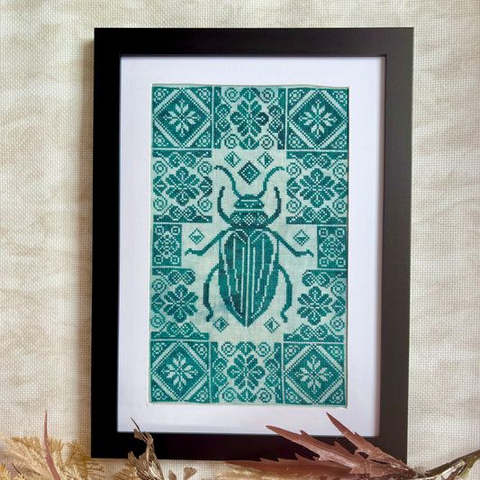 Framed cross-stitched pattern with a beetle design on a textured wall.