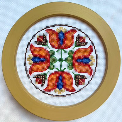 Circular cross-stitch pattern with orange flowers and green leaves in a wooden frame on a light blue background.
