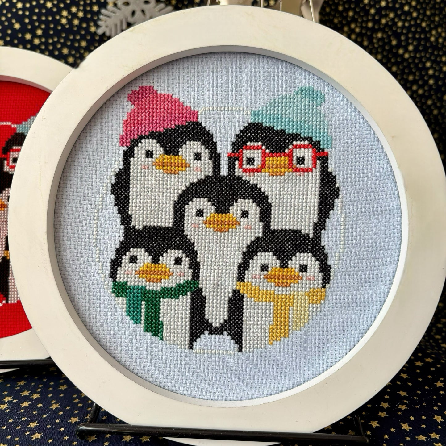 Cross-stitched penguins with accessories in a circular frame on a dark background