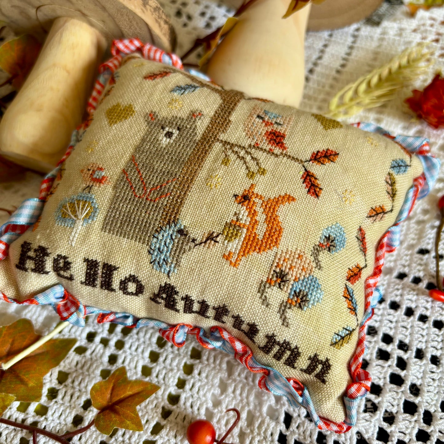 Handmade cross stitch pillow with woodland animals, colorful autumn leaves, and the text "Hello Autumn," stitched on linen and surrounded by fall decor