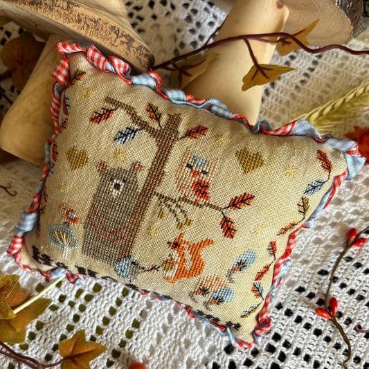 Handmade cross stitch pillow with woodland animals, colorful autumn leaves, and the text "Hello Autumn," stitched on linen and surrounded by fall decor