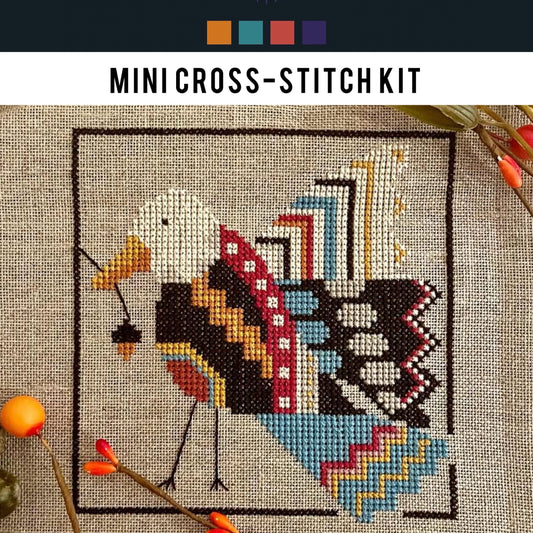 Mini cross-stitch kit with a completed design of a bird and acorn on fabric.