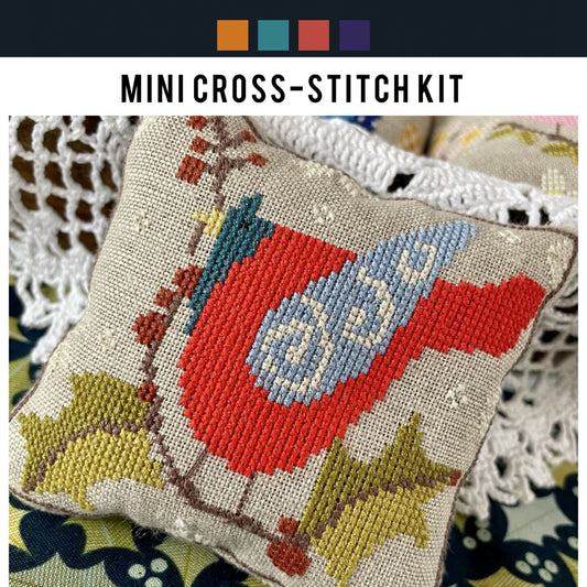Mini cross-stitch kit with a red and blue bird design on a textured fabric background.