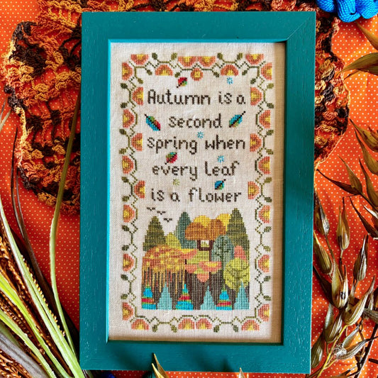 Framed cross-stitched artwork with autumn-themed text and colors on a decorative background.