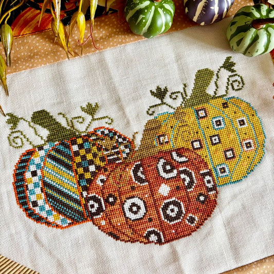 Cross-stitched pumpkins on fabric with decorative patterns, surrounded by small pumpkins.