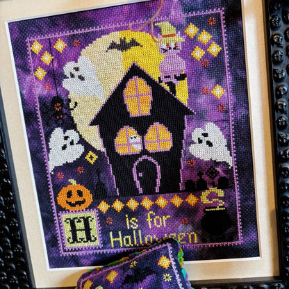 Cross-stitched Halloween design with a haunted house and pumpkins on purple fabric.