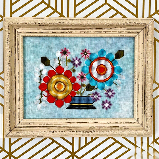 Framed cross-stitched floral design on a blue background with a decorative gold frame.